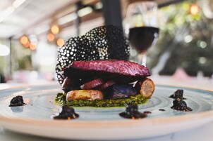 Un plato de comida con filete, verduras y salsa, con una copa de vino tinto al fondo.