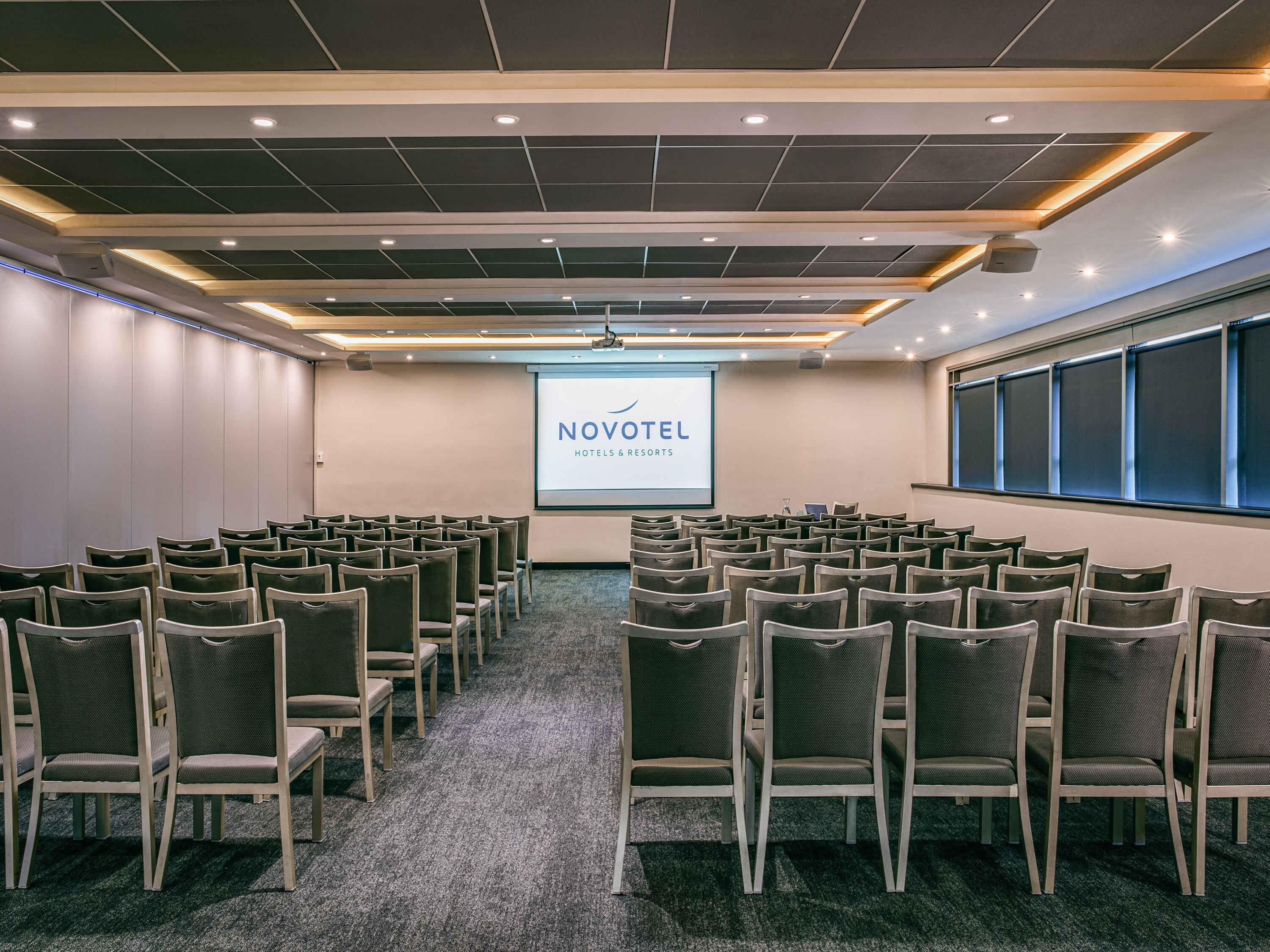 A conference room with rows of chairs, a projector screen displaying 'NOVOTEL HOTELS & RESORTS,' and a large window on the right.
