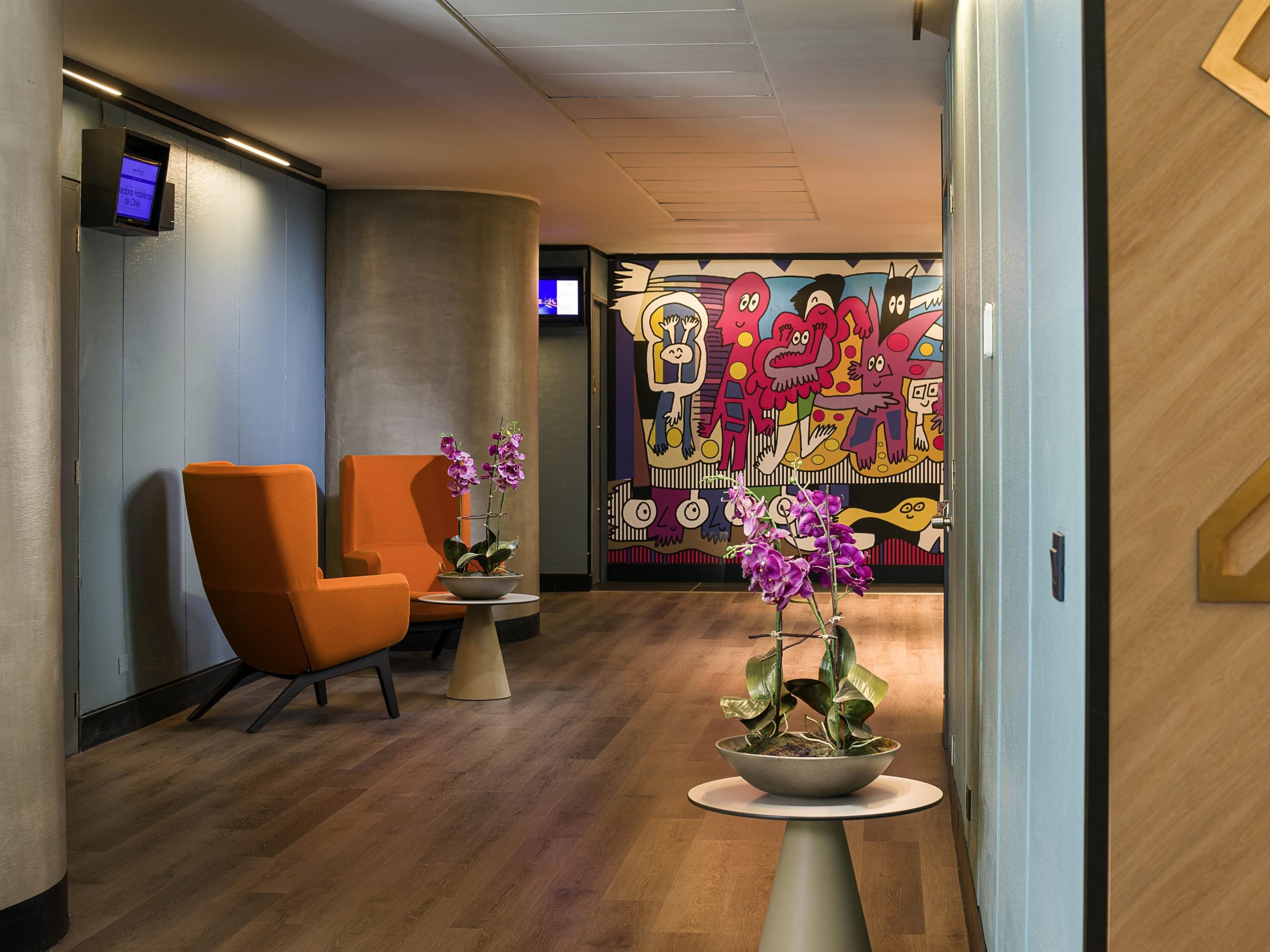 Um lobby moderno com duas cadeiras laranja, piso de madeira e um mural abstrato colorido na parede.