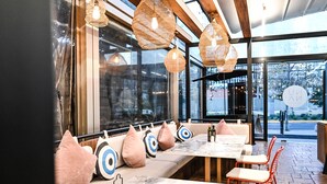 Een restaurantinterieur met een lange balie, zitplaatsen met kussens, hangende rieten lampen en grote ramen met uitzicht op de straat.