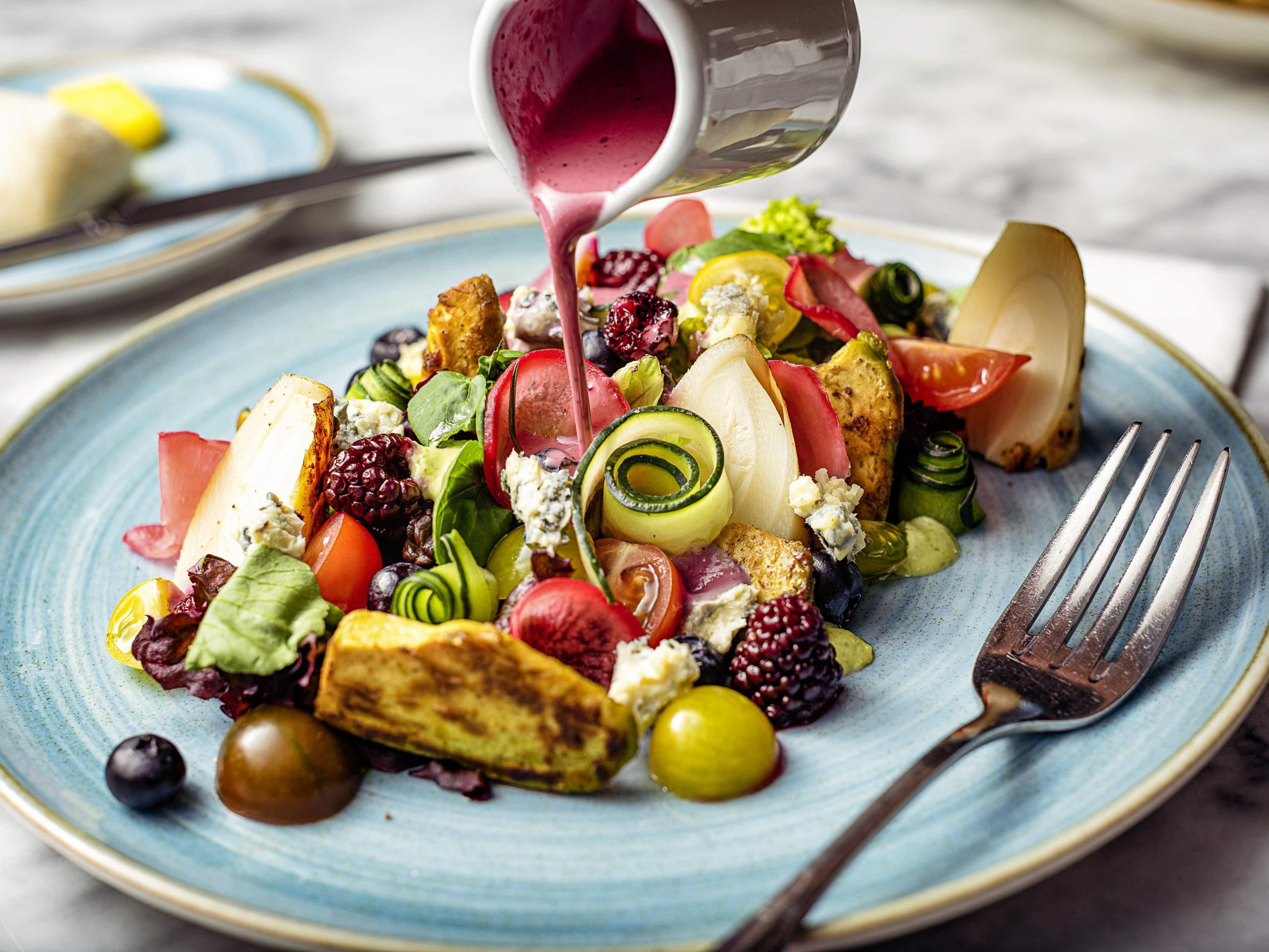 A plate of mixed greens, fruits, and cheese with a dressing being poured over it.