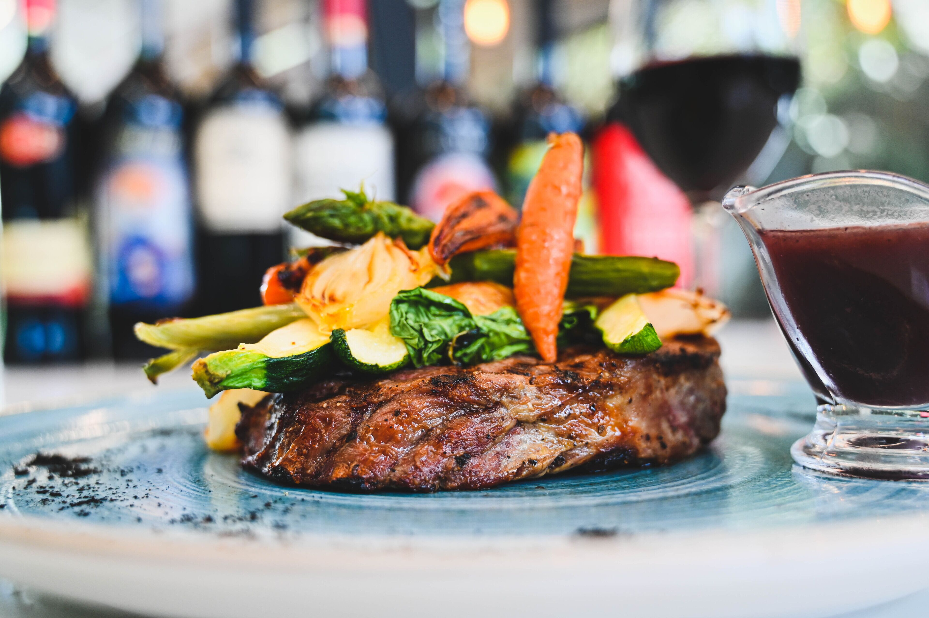 Ein Steak mit Gemüse auf einem Teller, ein Glas Rotwein und Flaschen im Hintergrund.