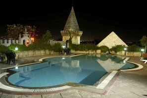 A swimming pool with a stone border, a pyramid-shaped structure, and a tower with a pointed roof in the background. - Amrutha Castle (Hyderabad)