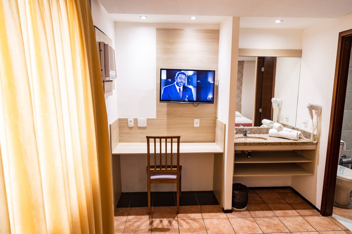 A hotel room with a TV mounted on the wall, a wooden chair, a small desk, a vanity with a mirror, and a shower area.