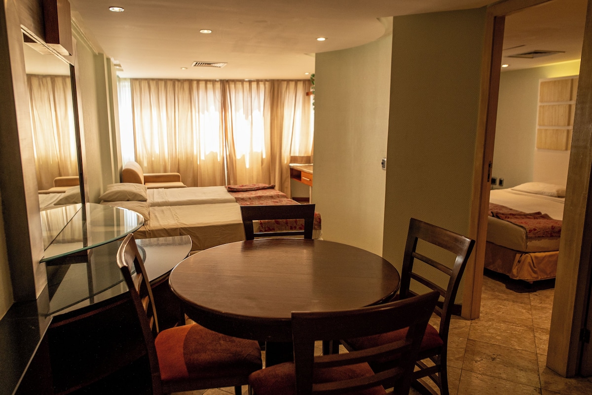 A hotel room with a glass dining table, two chairs, a sofa, a bed, and a bathroom.