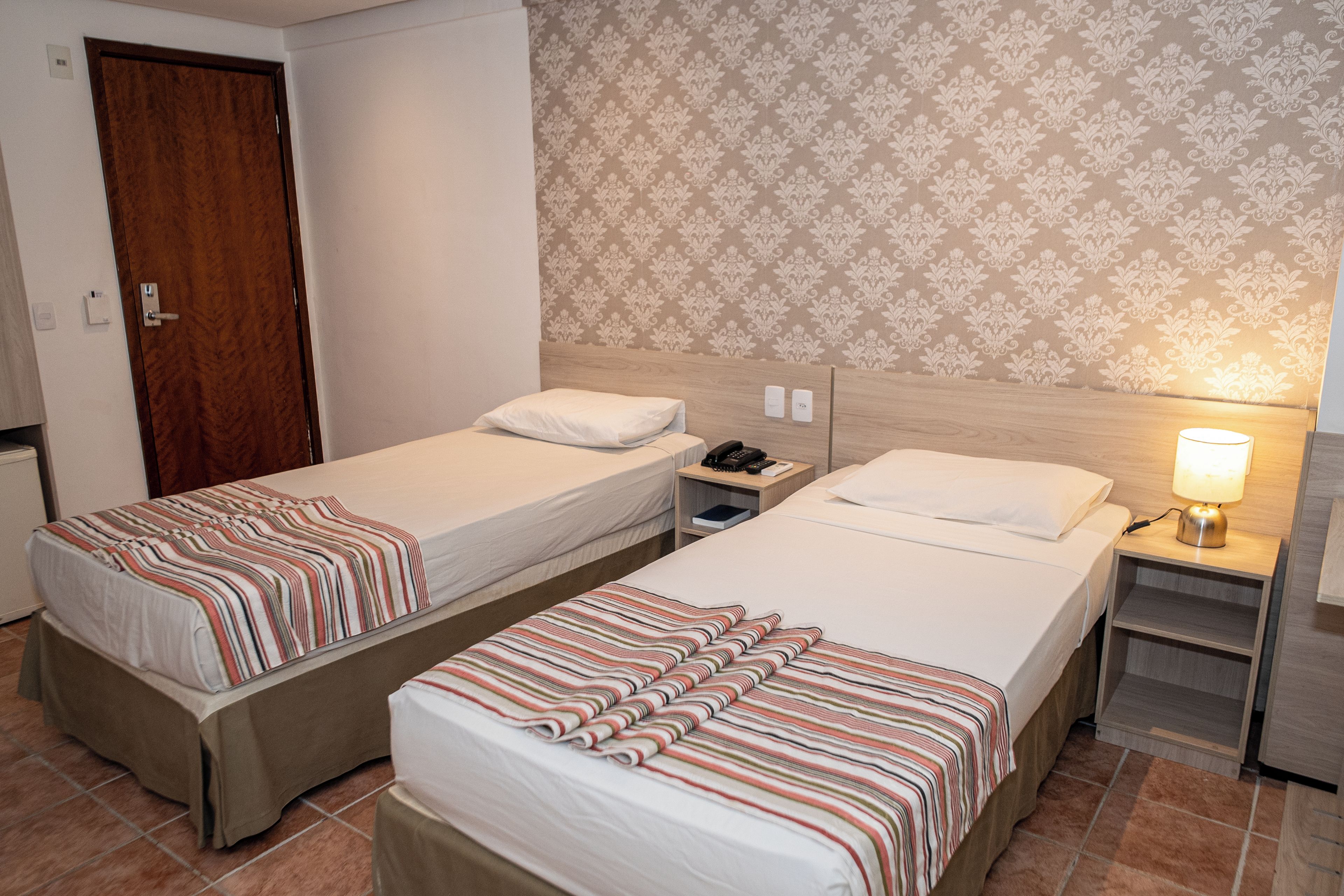 Une chambre d’hôtel avec deux lits simples, une tête de lit en bois, une table de chevet avec une lampe et un papier peint à motifs.
