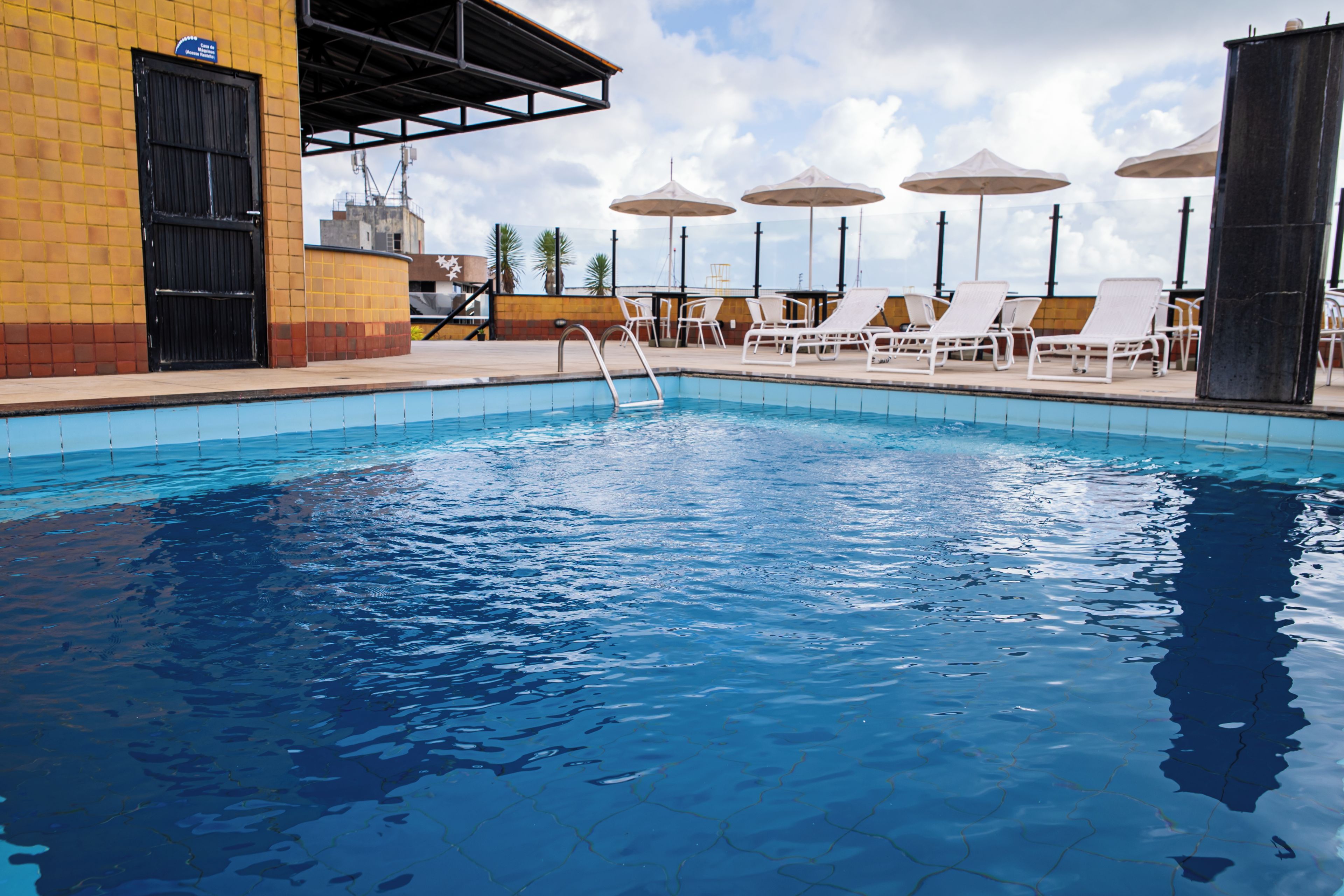 Una piscina con sillas de descanso y sombrillas, rodeada por un muro de baldosas y la entrada a un edificio.