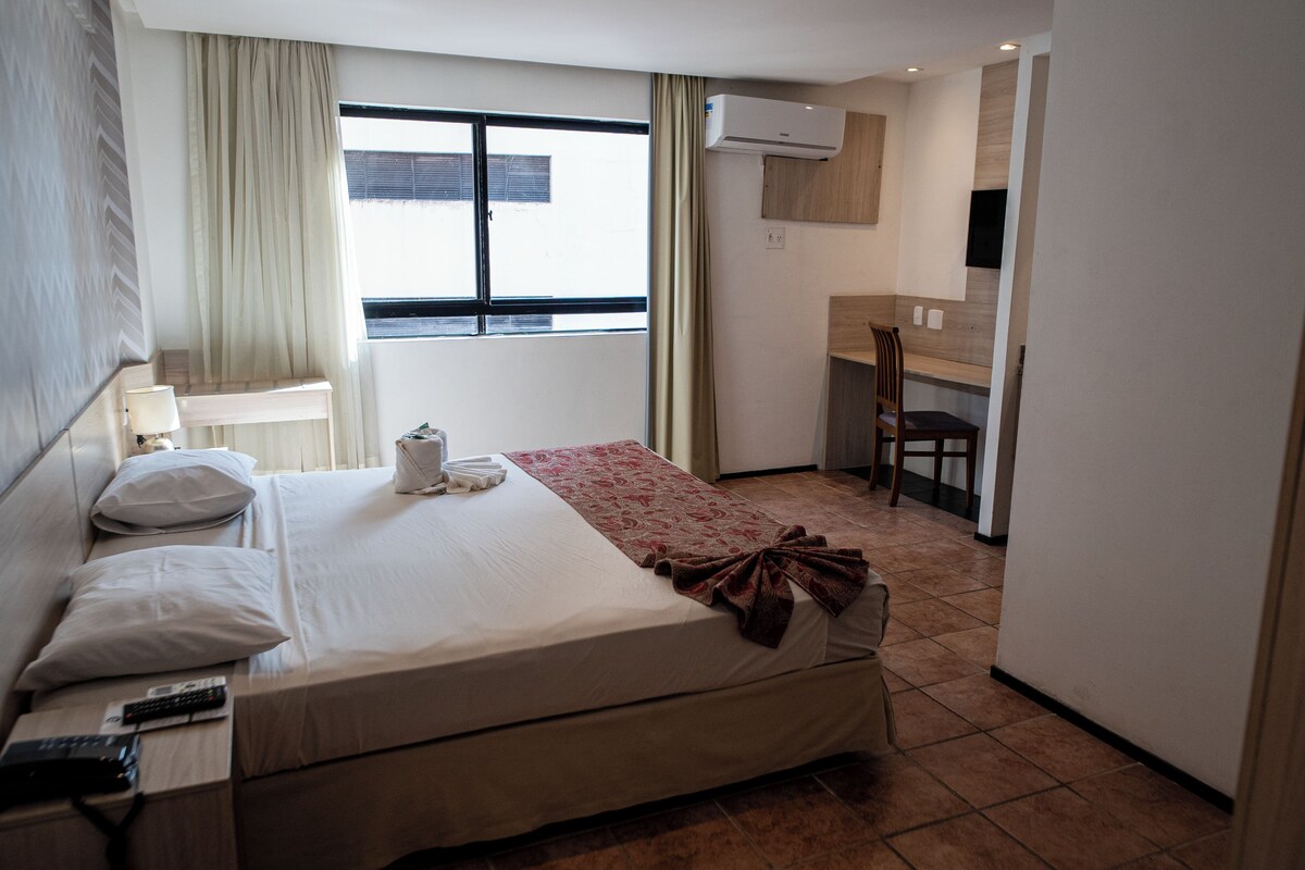 A hotel room with a bed, a desk, a chair, a window, and an air conditioning unit.