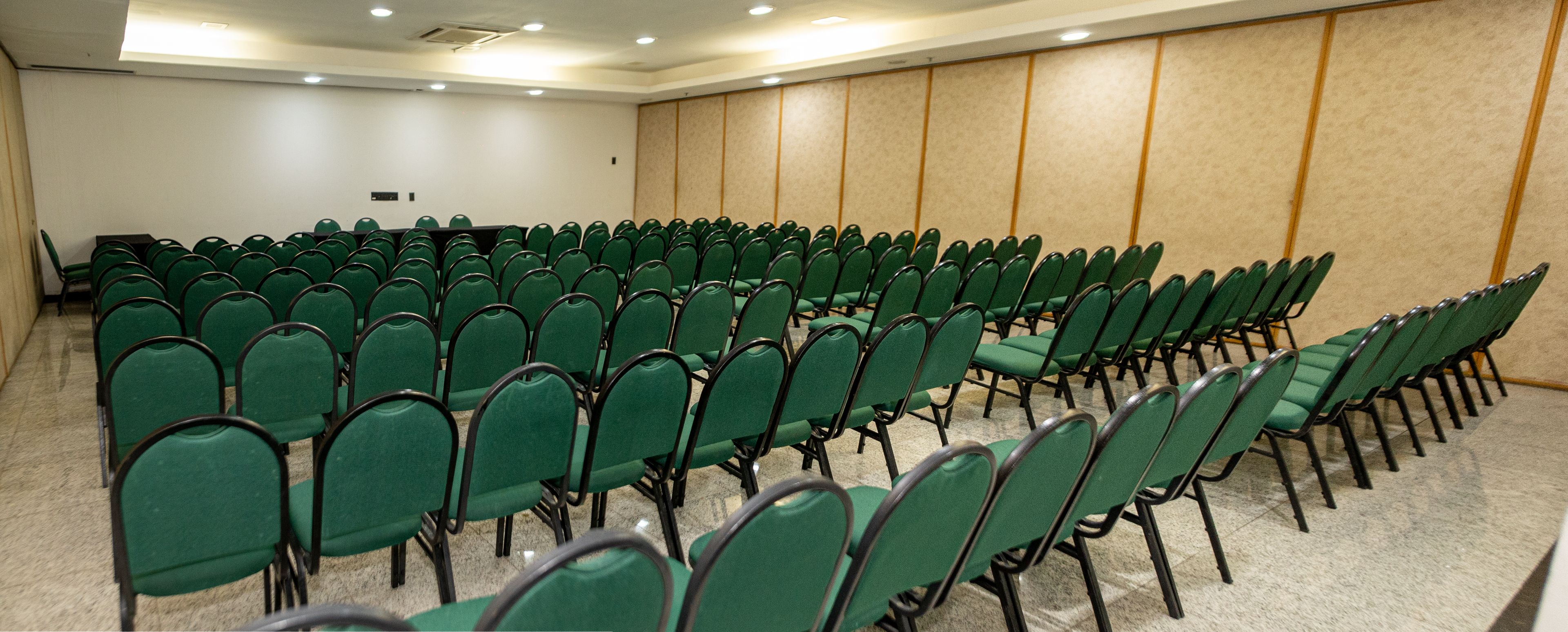 Sala de conferências com fileiras de cadeiras verdes e piso coberto por carpete.