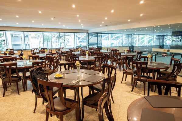 A spacious dining area with multiple tables and chairs, large windows offering views of the outside, and a modern ceiling design.