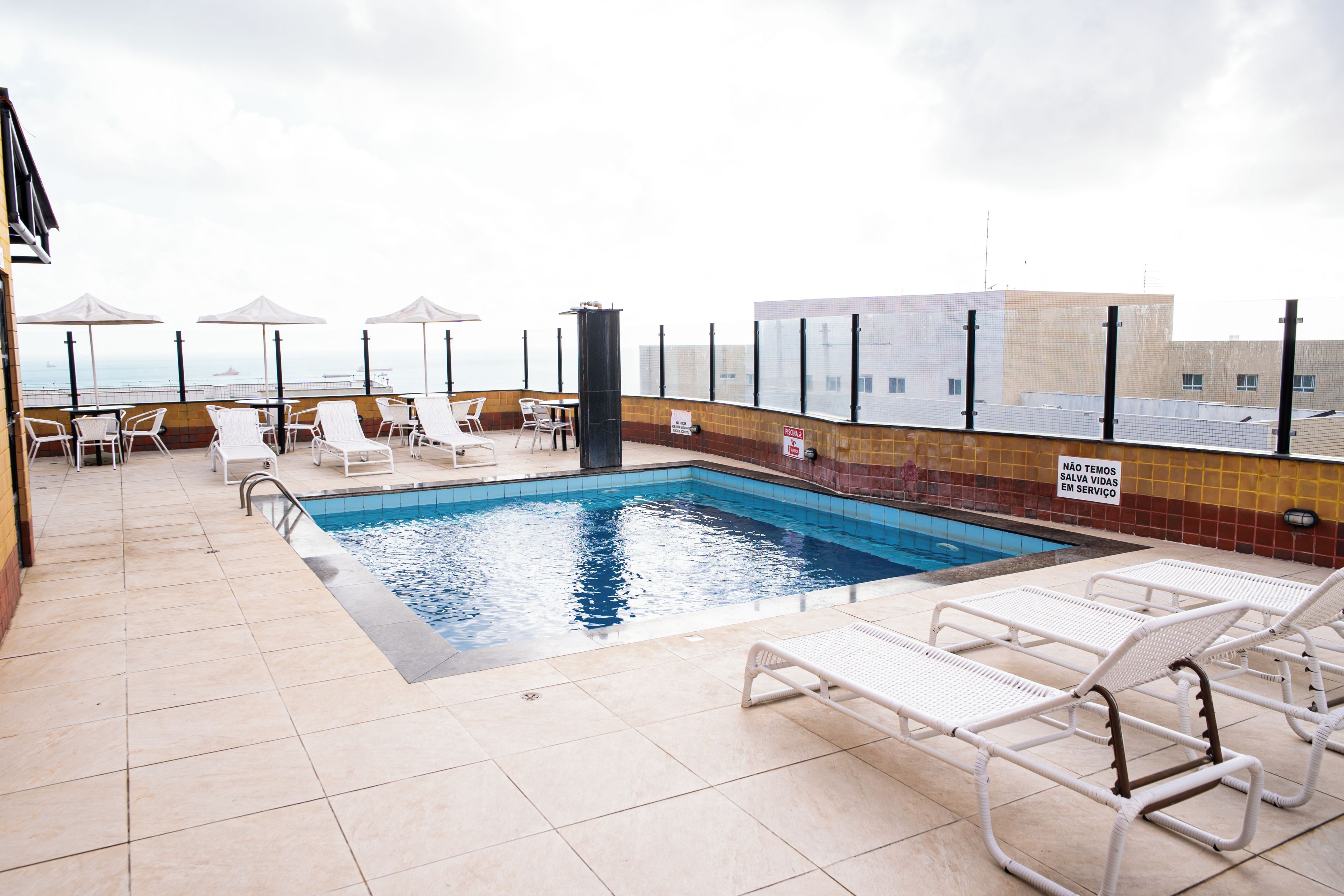 Un área de piscina en la azotea con sillones de descanso y sombrillas, vistas despejadas al océano y una piscina con baldosas.