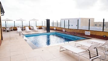 Un área de piscina en la azotea con sillones de descanso y sombrillas, vistas despejadas al océano y una piscina con baldosas.