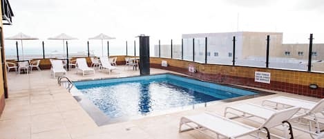 A rooftop pool area with lounge chairs and umbrellas, a clear view of the ocean, and a tiled pool.