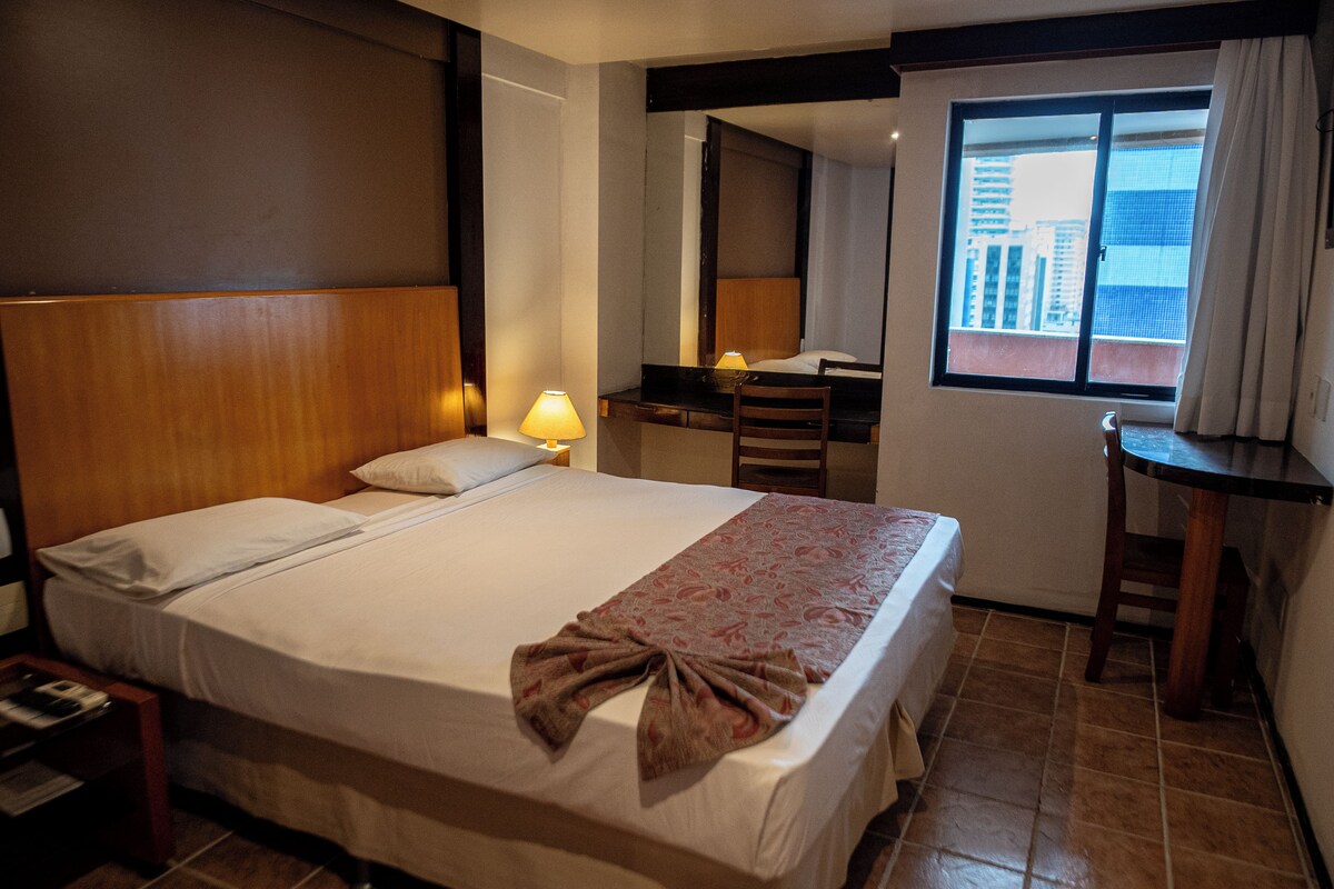 A hotel room with a bed, a desk, a chair, a lamp, and a window with a city view.