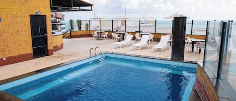 A swimming pool with lounge chairs and tables, a covered seating area, and a view of the sea.