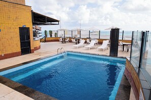 A swimming pool with lounge chairs and tables, a covered seating area, and a view of the sea. - Plaza Praia Suites (Fortaleza)