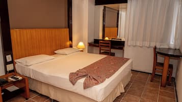 Quarto de hotel com cama, mesa de cabeceira, luminária e escrivaninha com cadeira.