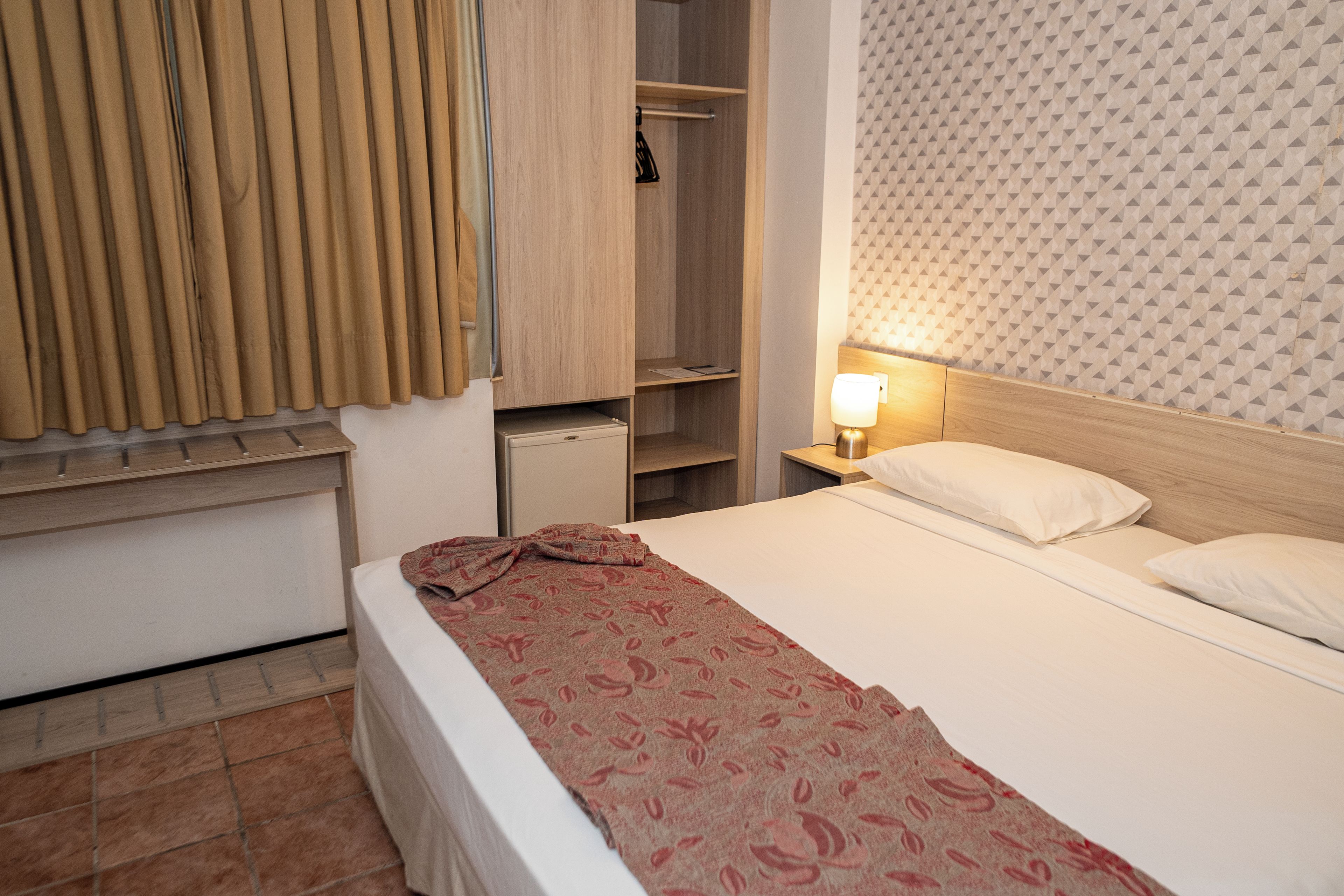 A hotel room with a bed, a nightstand with a lamp, a wardrobe, and a window with curtains.