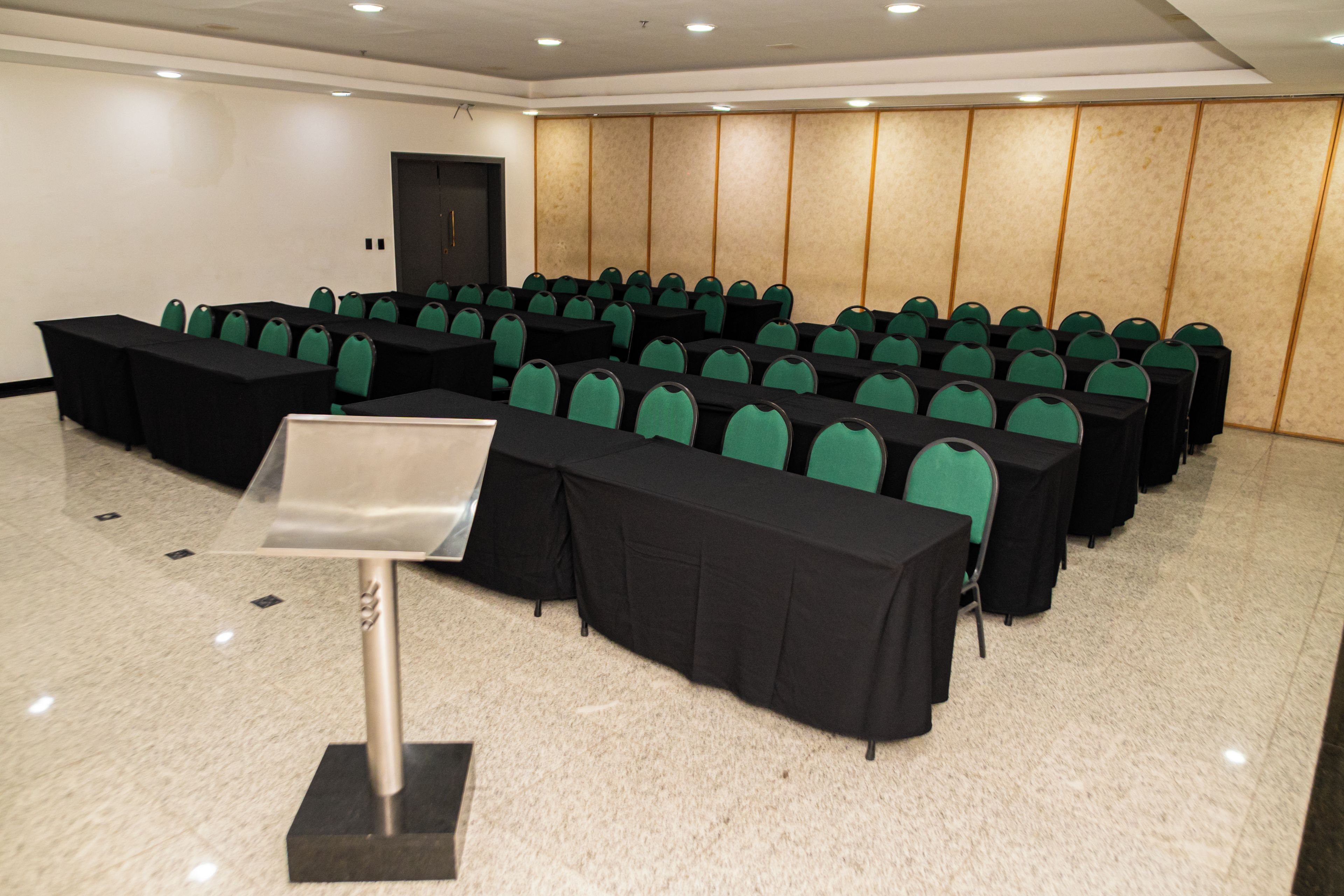 Sala de conferência com fileiras de mesas e cadeiras, um púlpito e uma porta.