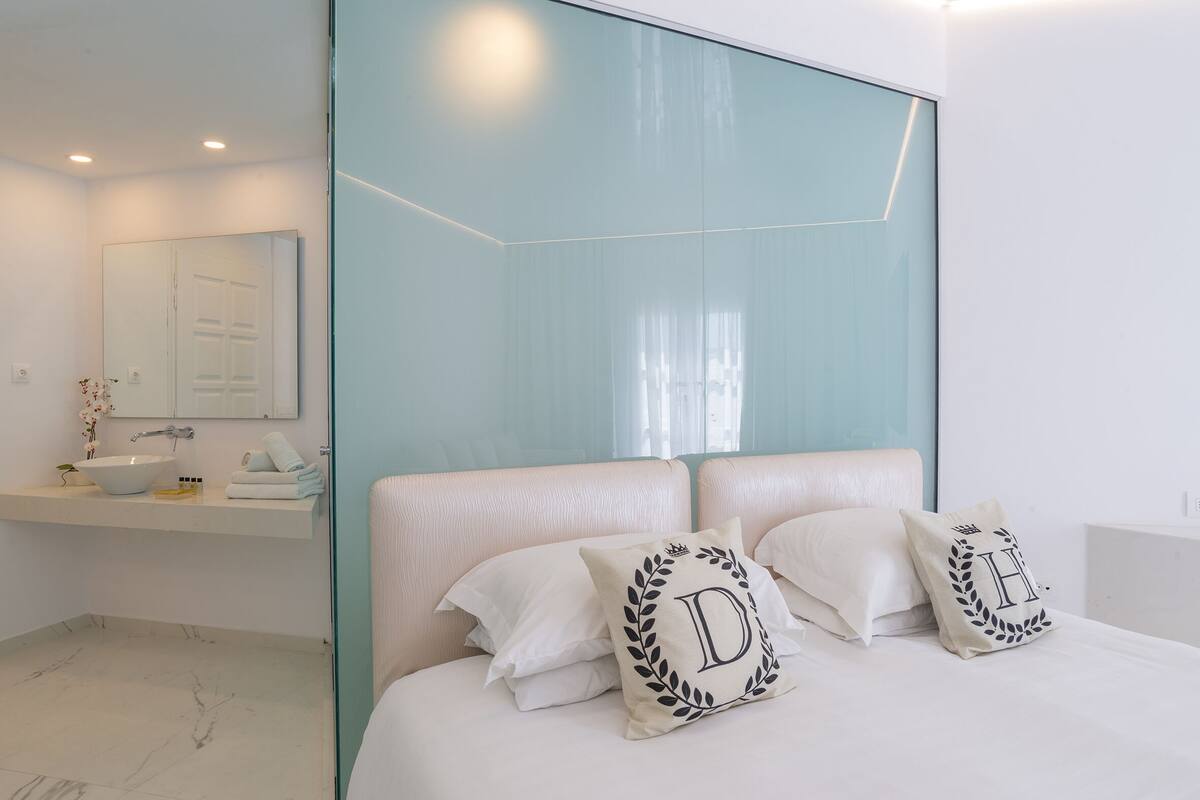 Un dormitorio moderno con una cama grande, dos almohadas con letras bordadas, una mesita de noche y un baño con lavamanos y espejo.