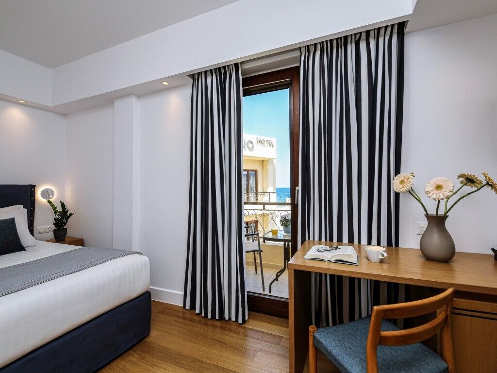 Une chambre d’hôtel avec un lit, un bureau et une chaise, un vase avec des fleurs et une vue sur la façade de l’hôtel.