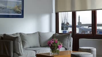 Ein Wohnzimmer mit grauem Sofa, einem Holztisch mit Blumenvase und Blick auf Boote und einen Leuchtturm durch große Fenster.