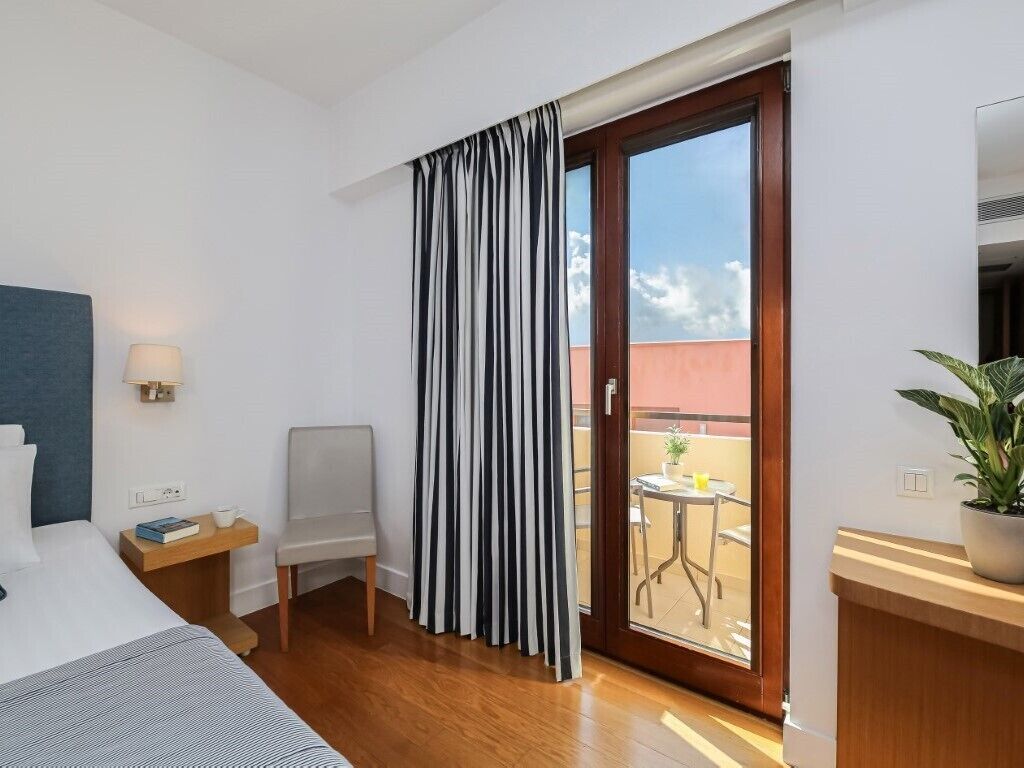 A hotel room with a bed, a chair, a desk, a potted plant, and a sliding glass door to a balcony.