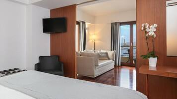 A hotel room with a bed, a TV, a sofa, a chair, a desk, and a potted plant.