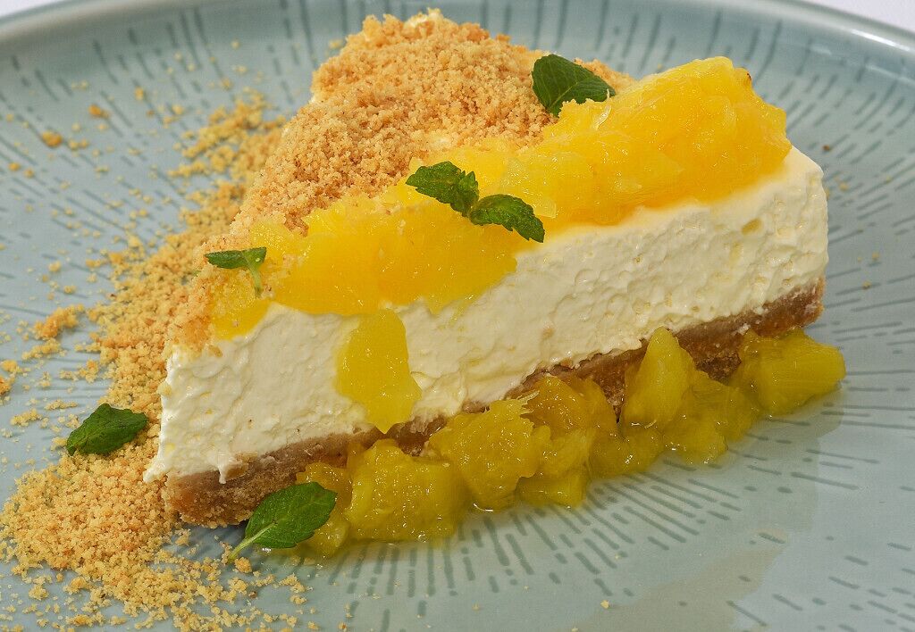 A slice of cheesecake with a crumbly topping and mango chunks, garnished with mint leaves.
