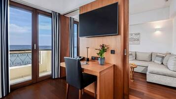 A room with a wooden desk, a flat-screen TV, a sofa, and a balcony with a view of the sea.