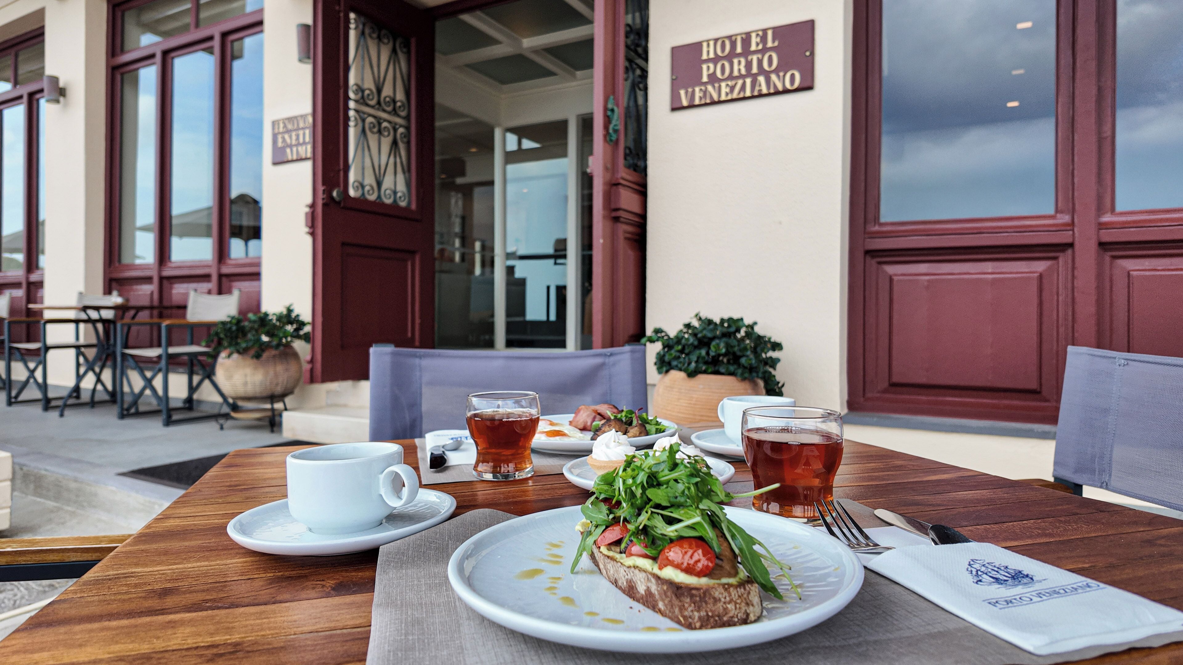 Une table à manger avec des assiettes de nourriture, une tasse et un verre de thé, placée à l’extérieur d’un bâtiment sur lequel on peut lire l’inscription « HÔTEL PORTO VENEZIANO ».