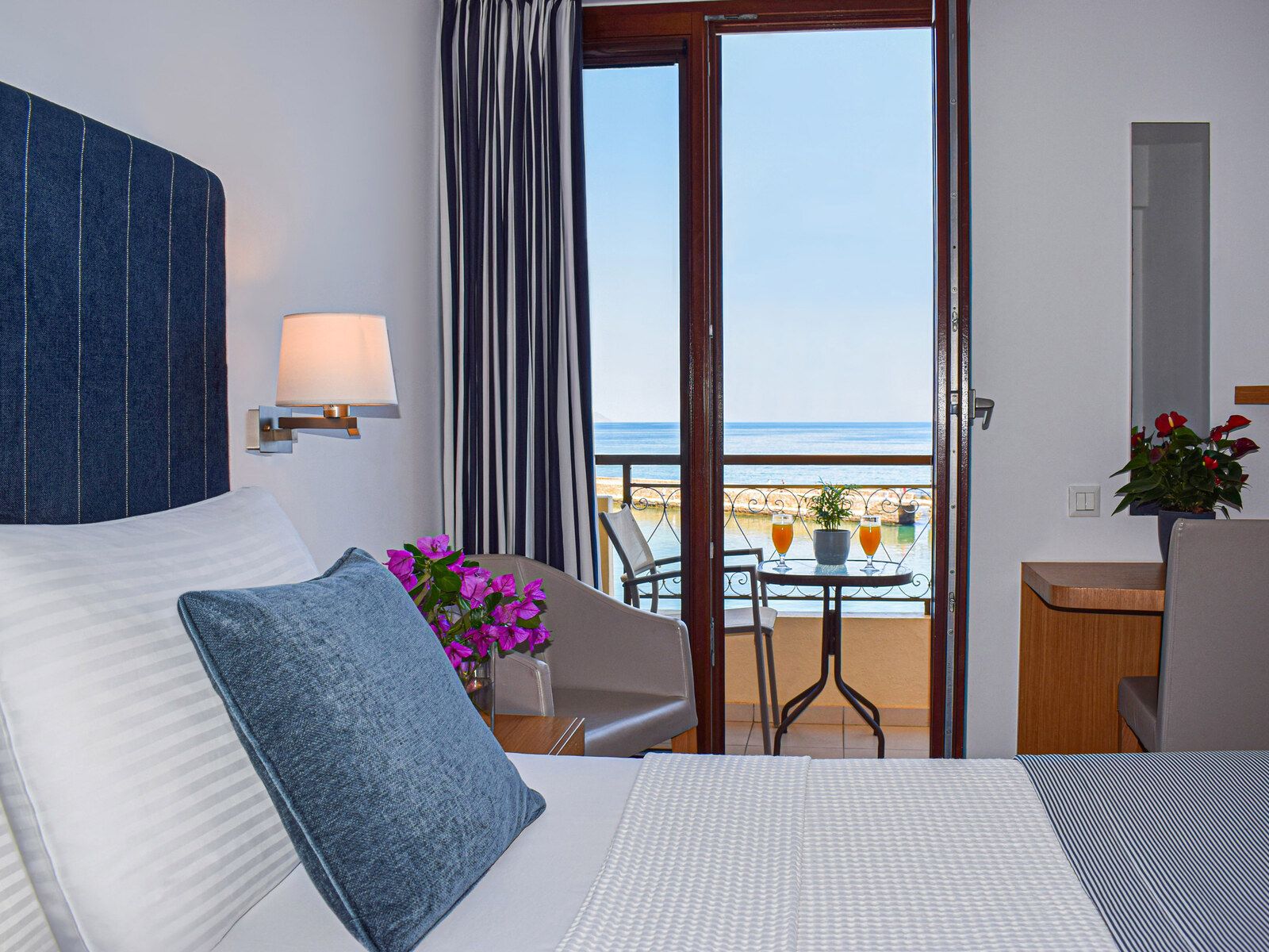 A hotel room with a bed, a sitting area with a table and chairs, a view of the beach, and a balcony with a table and chairs.