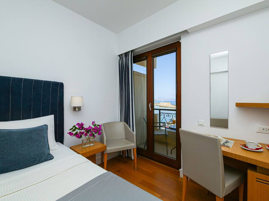 A hotel room with a bed, a desk, a chair, a small table with a lamp, and a view of the sea through a glass door.