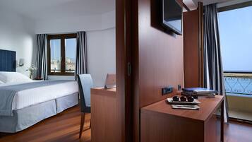 Un cuarto de hotel con una cama, un escritorio con una computadora portátil, un televisor y un balcón con vista al mar.