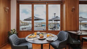 A dining area with a table set for breakfast, large windows with a view of a harbor, and wooden paneling. - Porto Veneziano Hotel (Chania)