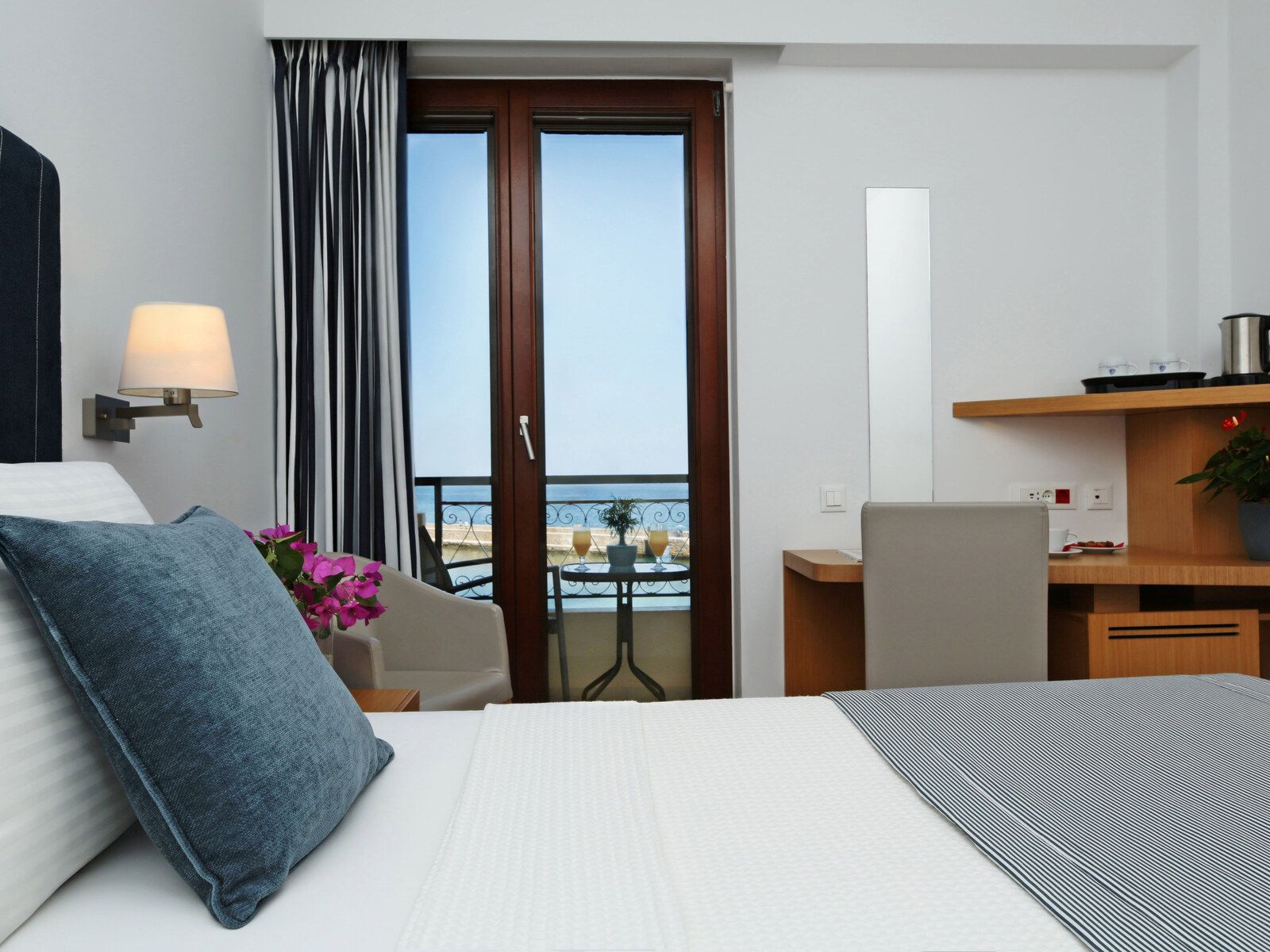 A hotel room with a bed, a desk, a chair, a balcony with a view, and a lamp.