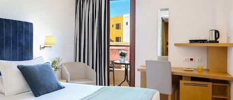 A hotel room with a bed, a desk, a chair, a small table with a plant, and a view of buildings outside.