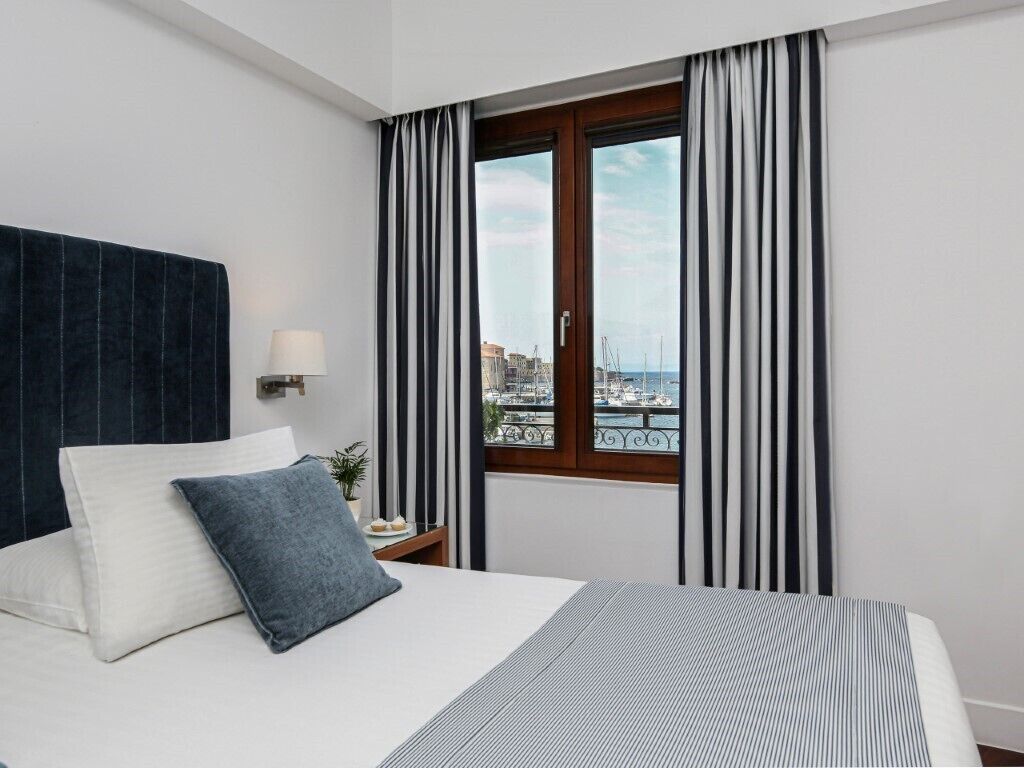 A hotel room with a bed, a nightstand, a lamp, a view of the city through a window with curtains.