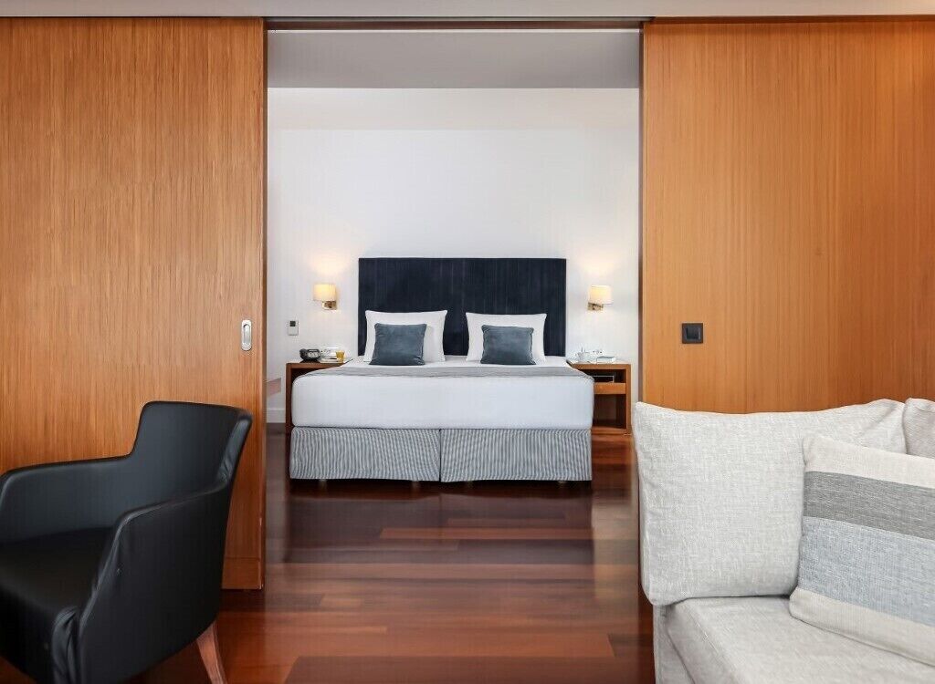 A hotel room with a bed, a black chair, a white sofa, and wooden paneling.