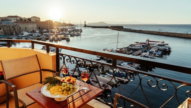 A balcony with a table set for a meal, overlooking a marina with boats and a distant view of a historic building. - Porto Veneziano Hotel (Chania)