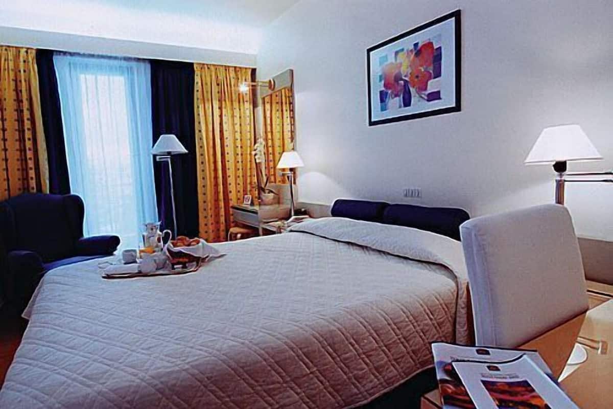 A hotel room with a large bed, a desk, two chairs, a lamp, and a painting on the wall.
