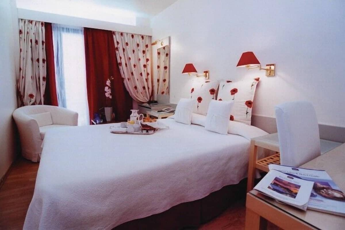 A hotel room with a large bed, two bedside lamps, a desk with a chair, and a window with curtains.