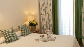 Una cama bien hecha con almohadillas a cuadros, una mesita de noche con una lámpara, una bandeja con tazas y una jarrilla, y una ventana con cortinas verdes.