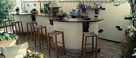 Un espace bar extérieur doté d’un sol carrelé, de tabourets en bois, d’une nappe à carreaux et de plantes en pot.