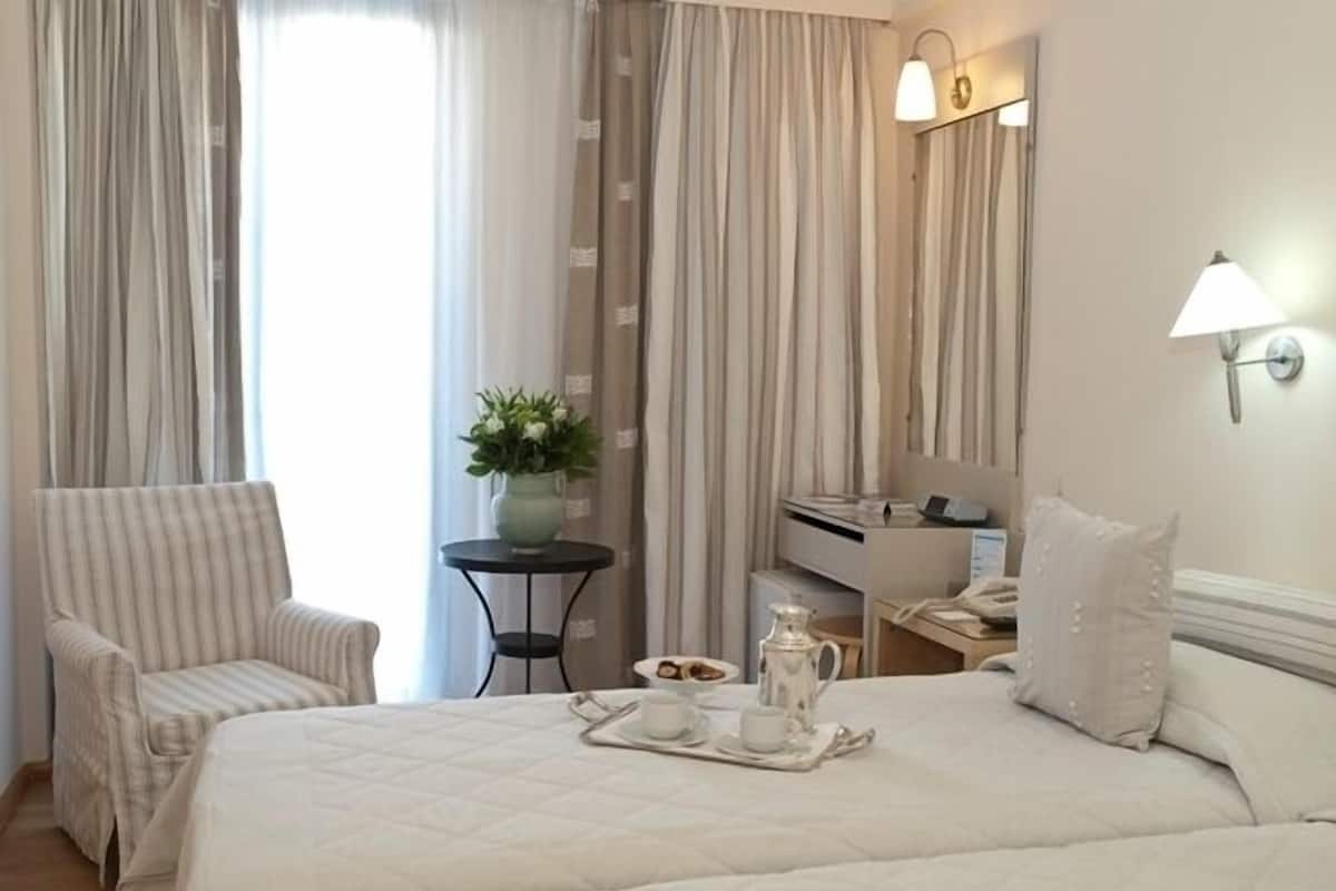 A hotel room with two beds, a chair, a small table with a tray of breakfast items, a desk with a lamp, and curtains.