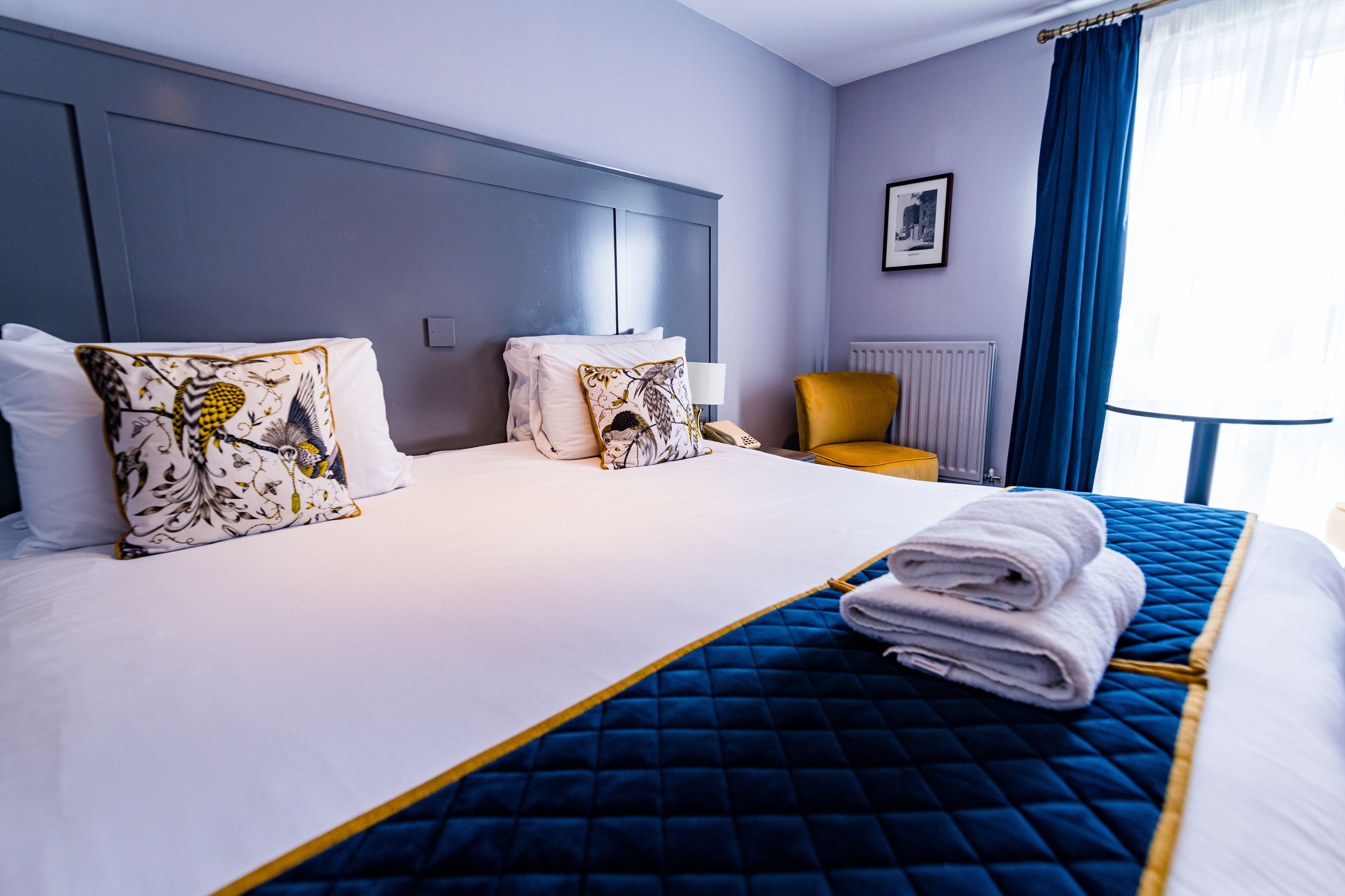 A hotel room with a large bed, two decorative pillows, a blue quilted bedspread, a folded towel, a chair, and a window with blue curtains.