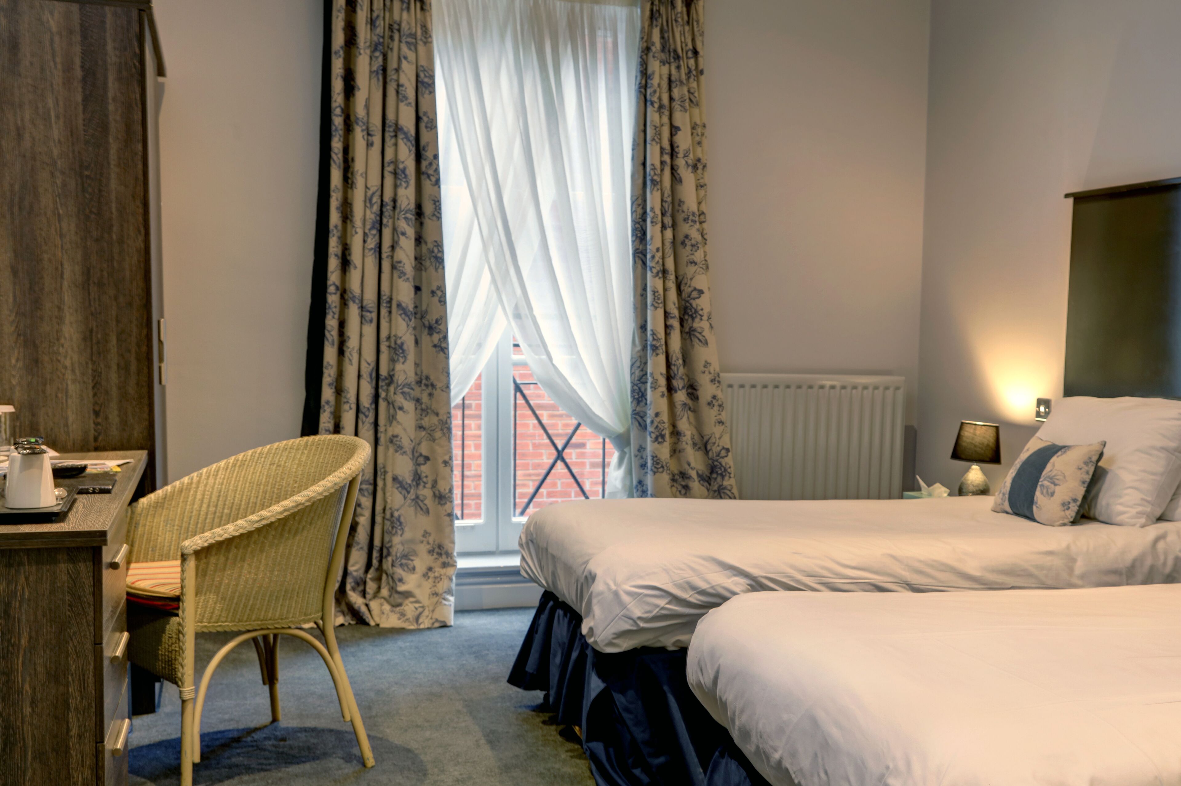 A hotel room with a bed, a desk with a chair, a window with curtains, and a lamp on the bedside table.