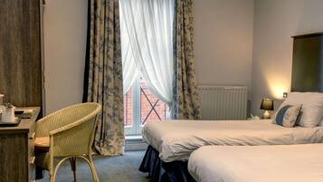 A hotel room with a bed, a desk with a chair, a window with curtains, and a lamp on the bedside table.