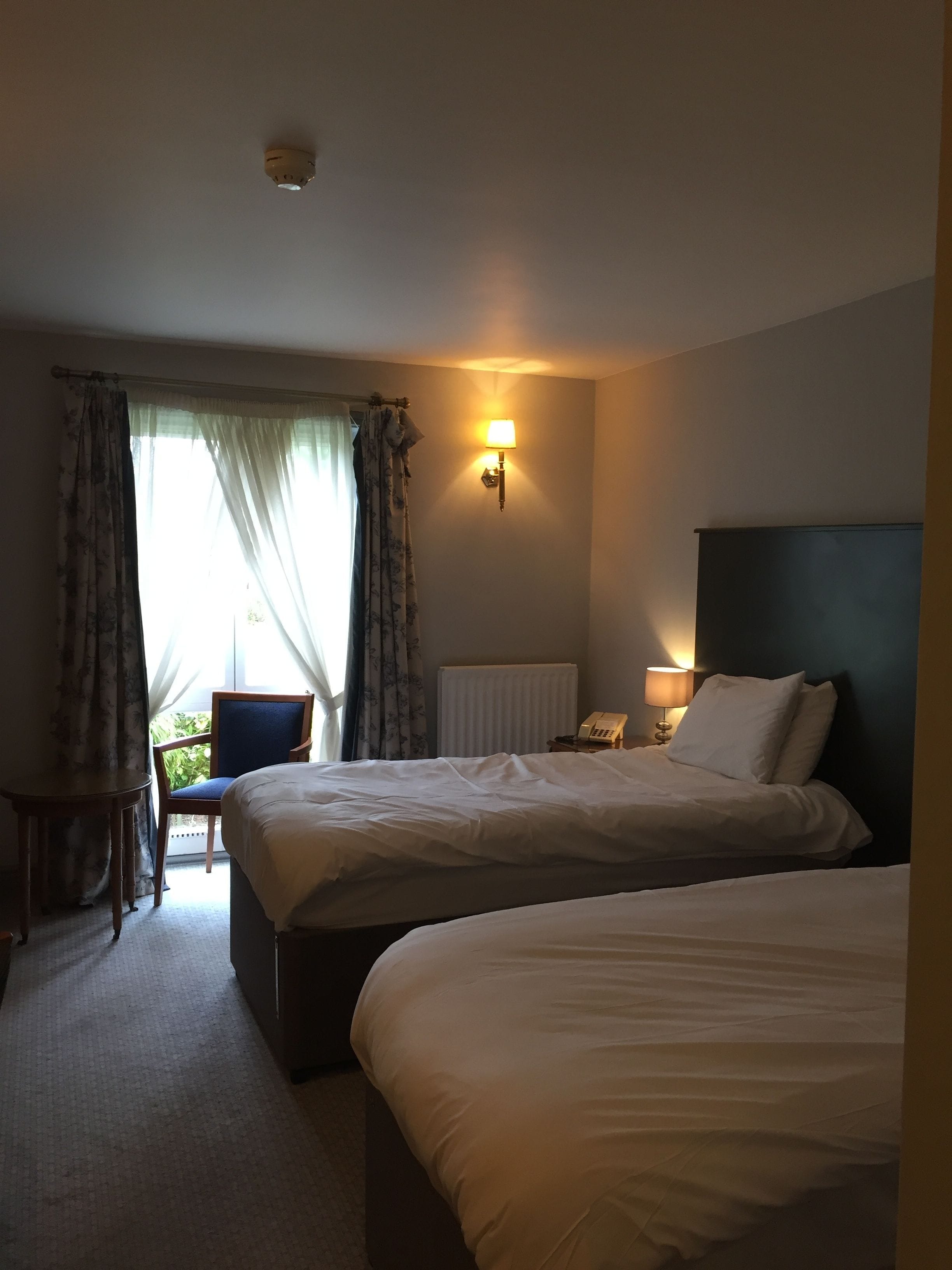 Habitación de hotel con dos camas, una silla, una mesa, una lámpara y una ventana con cortinas.