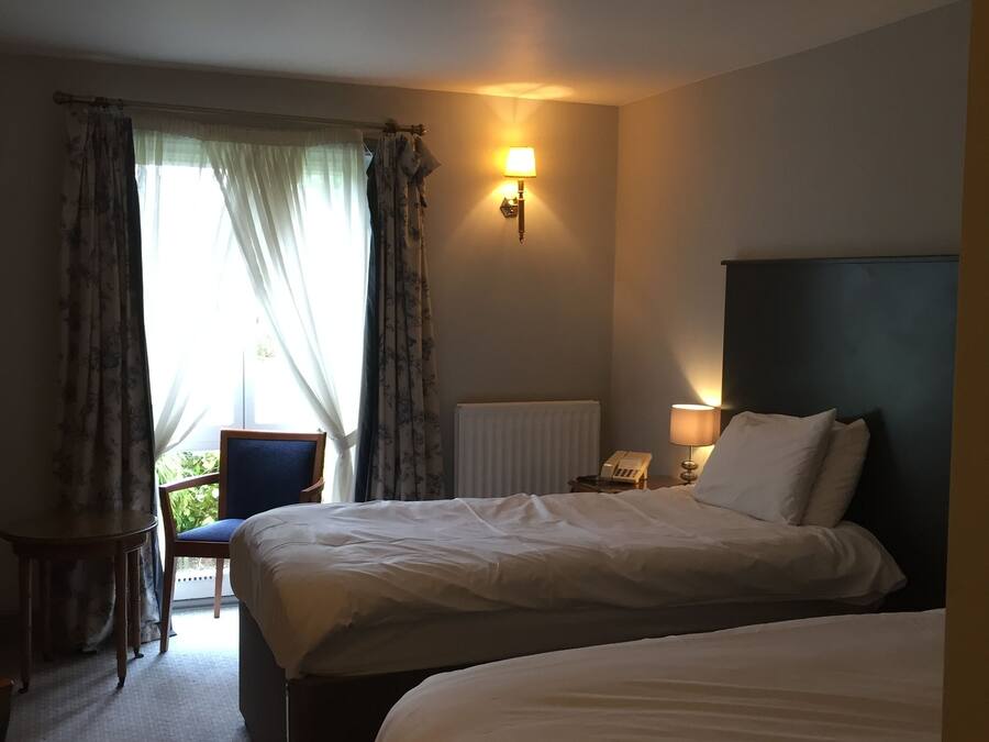 Habitación de hotel con dos camas, una silla, una mesa, una lámpara y una ventana con cortinas.